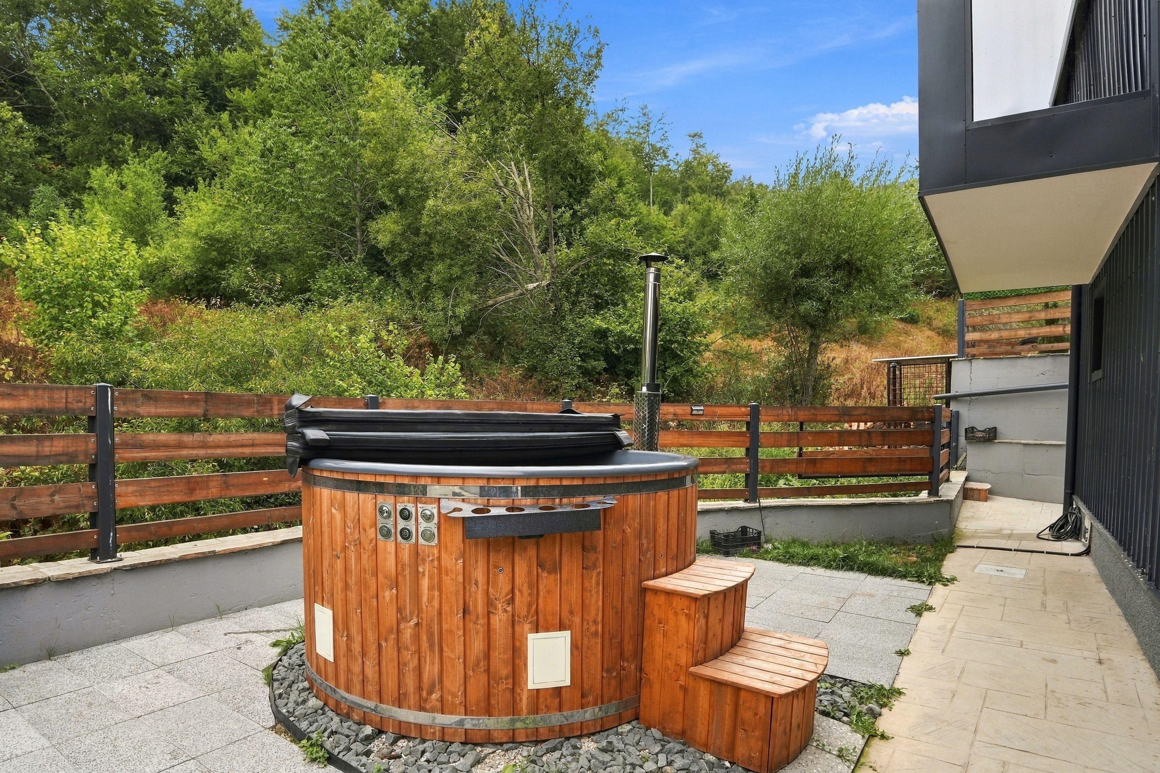 A wooden hot tub with a metal heater, surrounded by a wooden fence and a bench, set against a backdrop of trees and a building.