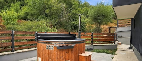 A wooden hot tub with a metal heater, surrounded by a wooden fence and a bench, set against a backdrop of trees and a building.