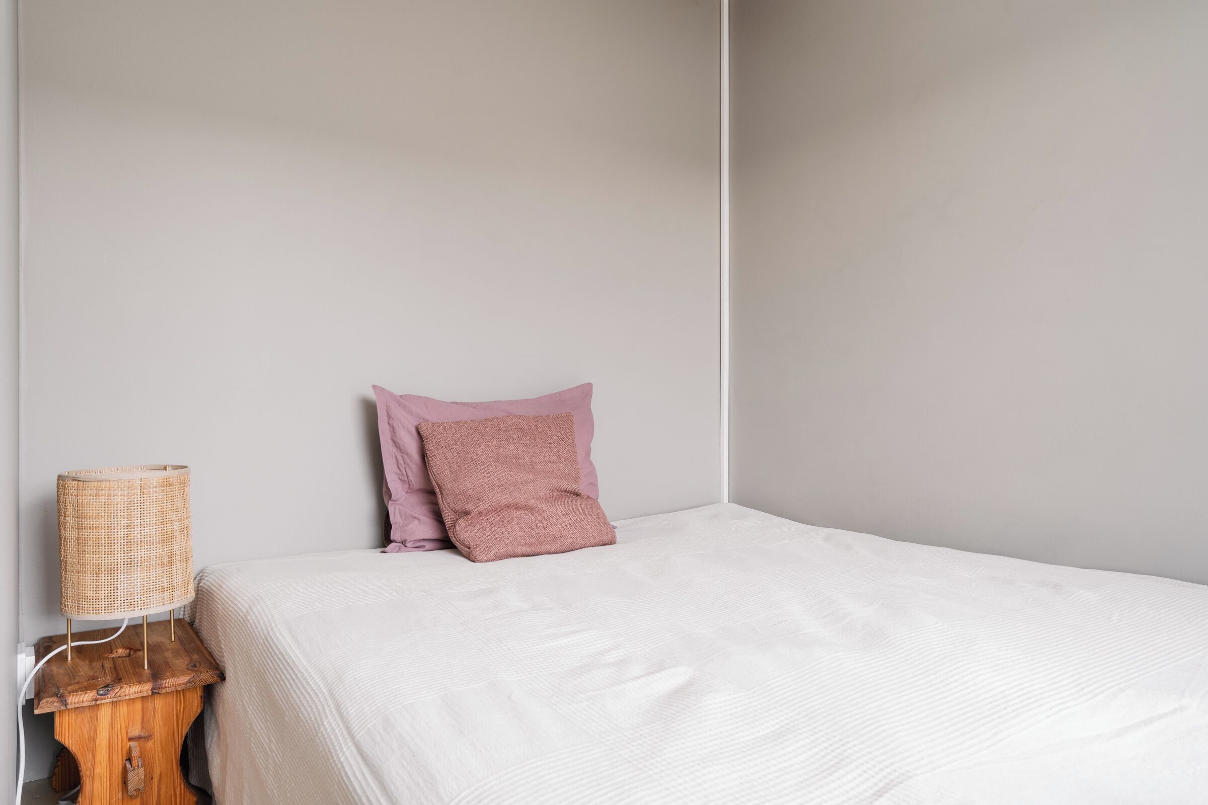 Una cama bien tendida con sábana blanca y almohada rosa, una mesita de noche de madera con lámpara de fibra y una pared lisa.
