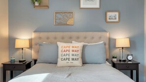 En pænt redt seng med en pude, der står "CAPE MAY EST. 1848" og en betrukket sengegavl. To sengeborde, hver med en lampe og en lille plante.
