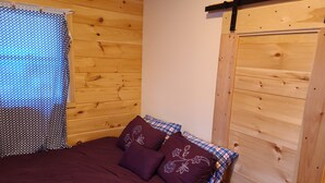 Ein Zimmer mit Holzboden, einem Bett, einem Fenster mit gemustertem Vorhang und einer Holztür.