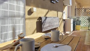 A breakfast table with two mugs, a plate, and a spoon. A towel hangs on the wall, and a window with a mesh screen is visible.