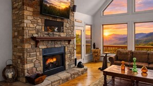 Un camino in pietra con una TV, un tavolo di legno con oggetti decorativi e vista sulle montagne attraverso ampie finestre.