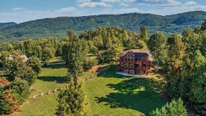 Un'ampia casa a due piani in legno con veranda che abbraccia l'intera struttura, immersa nel verde e con dolci colline sullo sfondo.