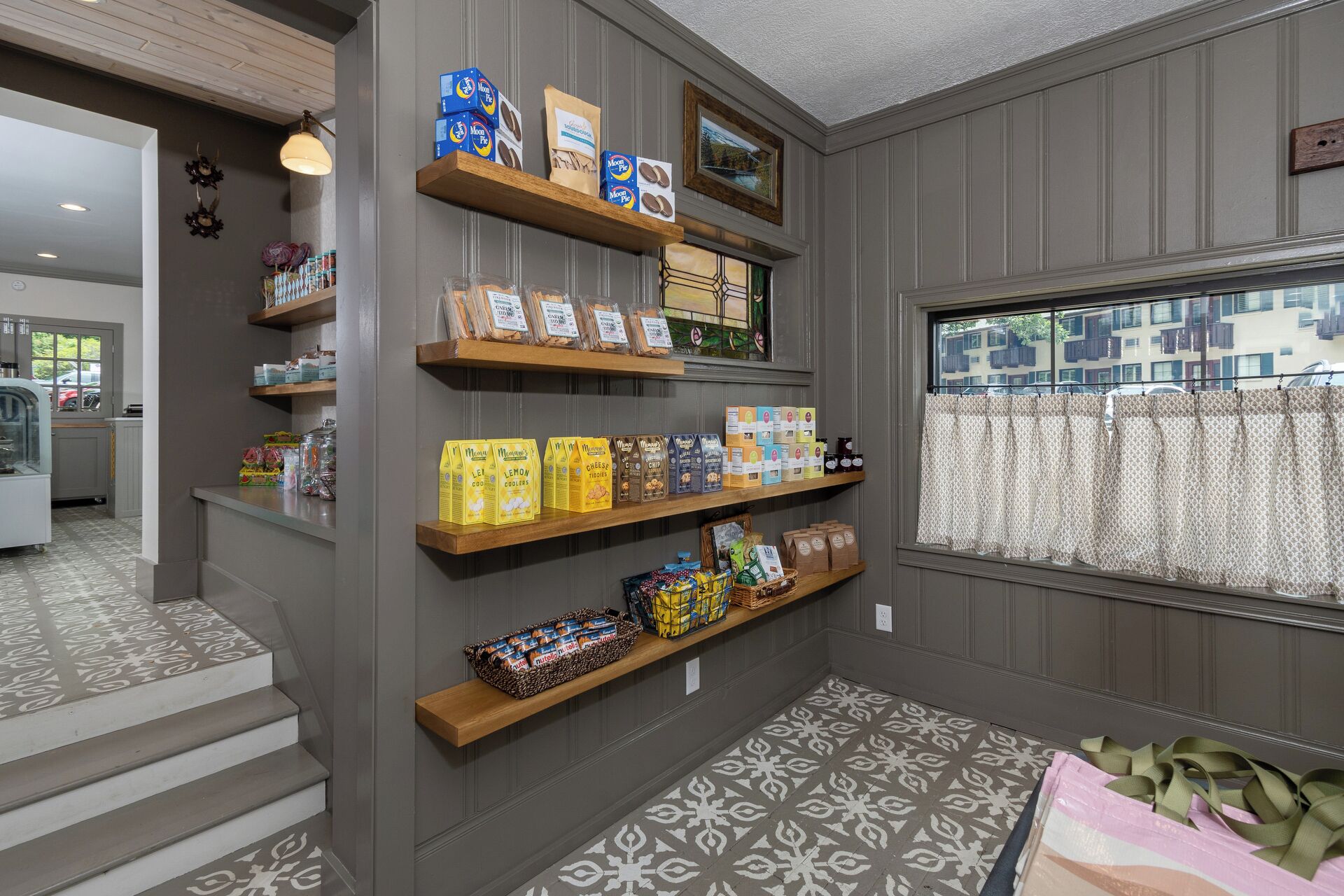 A store interior with shelves stocked with various packaged goods, a window with patterned curtains, and a view of a residential area outside.
