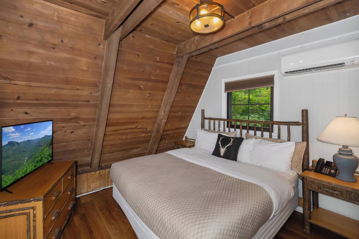 A bedroom with a wooden ceiling, a bed with white bedding, a nightstand with a lamp, and a television on a wooden dresser.