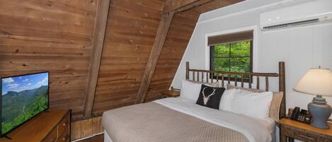Un dormitorio con techo de madera, una cama con ropa de cama blanca, una mesita de noche con lámpara y un televisor sobre un aparador de madera.