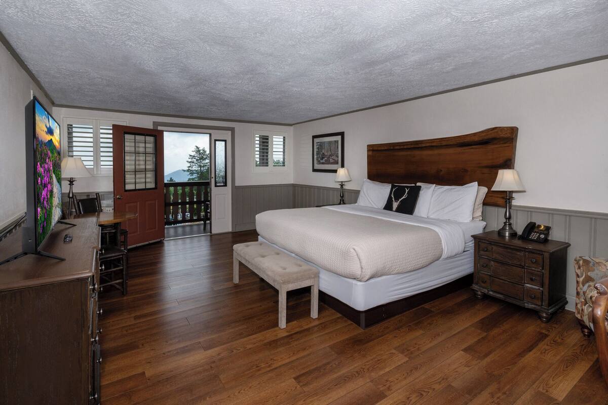 A bedroom with a large bed, wooden floor, a bench, a dresser, and a painting on the wall.