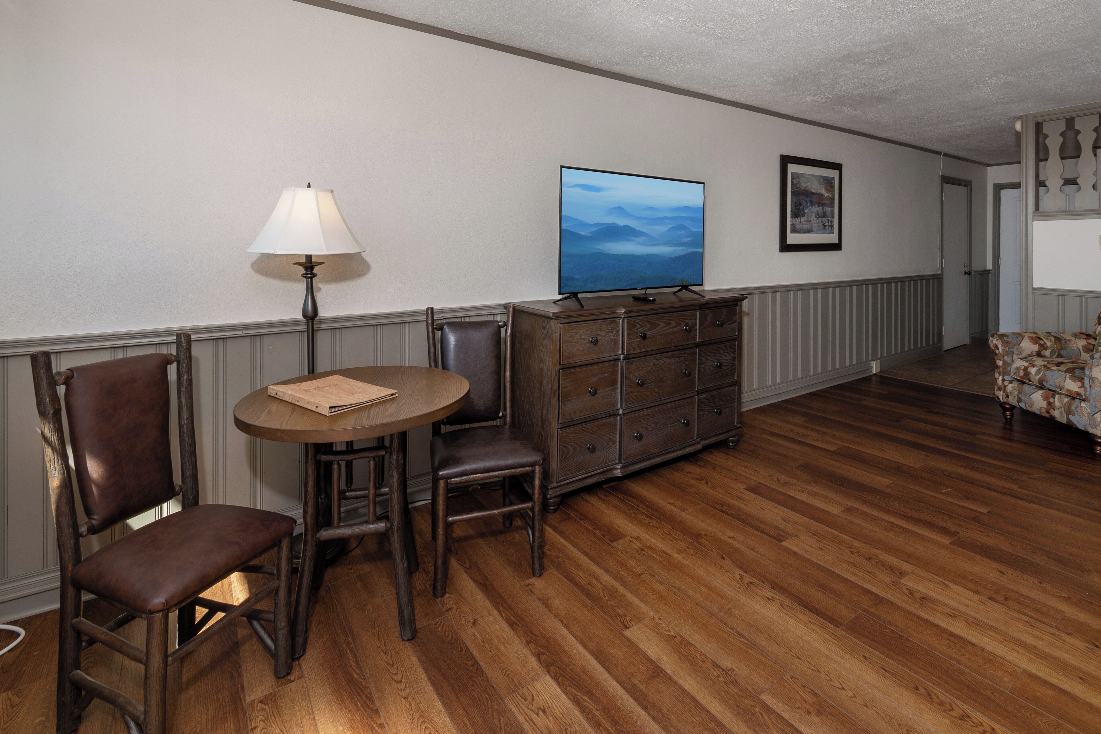 Une pièce avec un plancher en bois, une table ronde avec une lampe, un téléviseur et une commode.