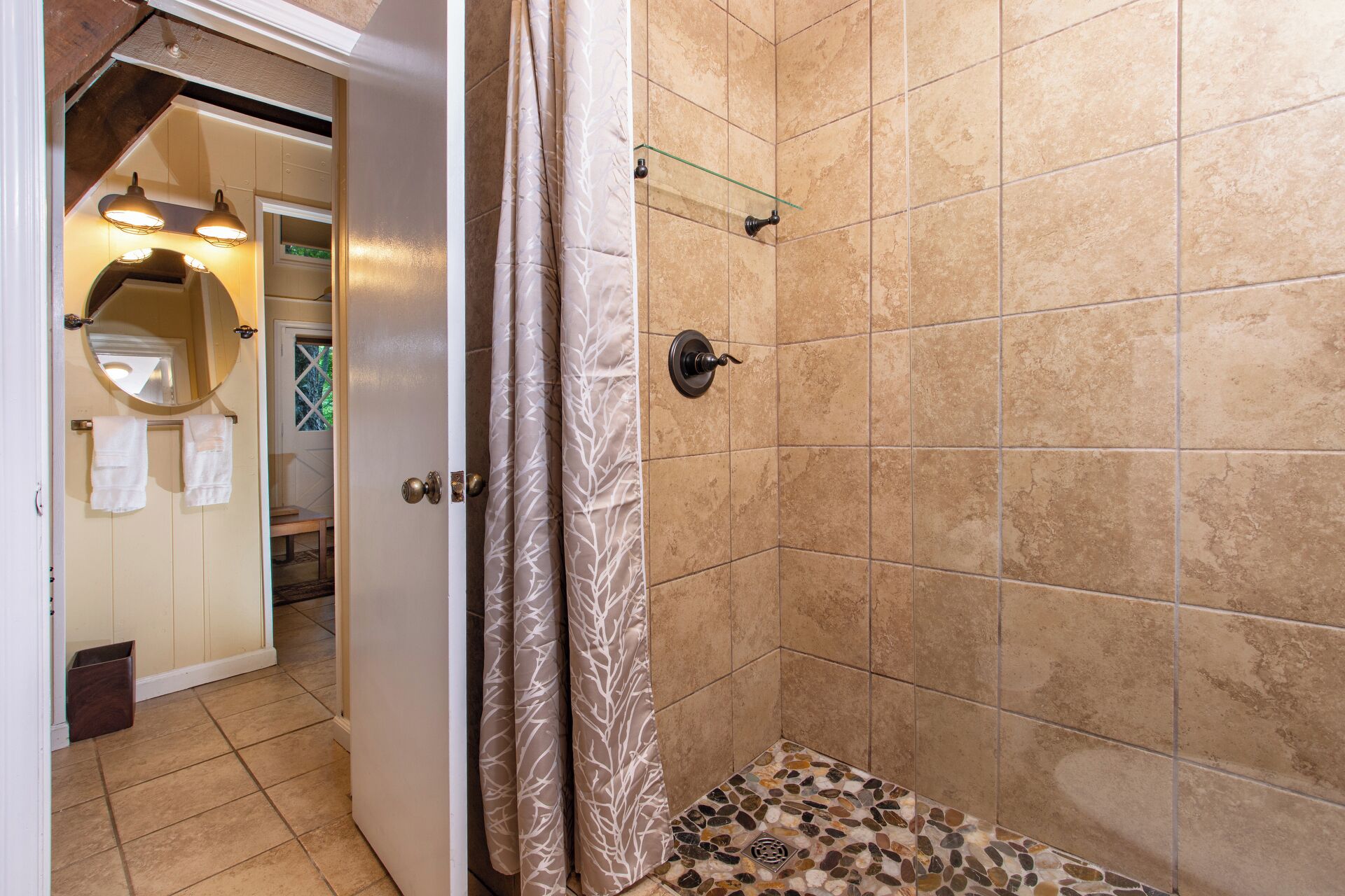 A bathroom with beige tiles, a shower with a glass shelf, and a door leading to a hallway.