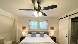Une chambre à coucher comprenant un lit, deux tables de chevet, un ventilateur de plafond, une fenêtre et une porte coulissante.