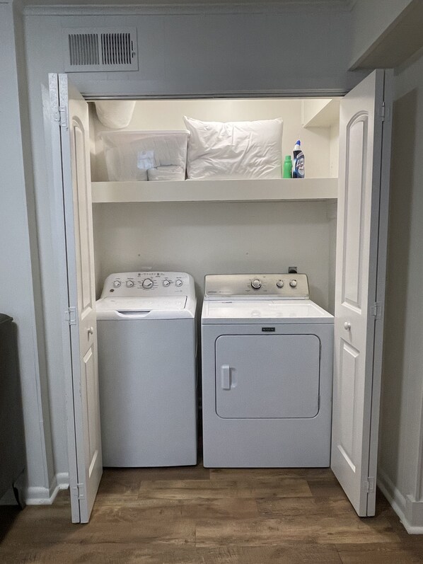 A laundry room with two washing machines and a dryer, with a shelf above containing a pillow and cleaning supplies. - Chic Converted Barn w/ 2 BR & 2.5 BA in Covington, LA (Covington)