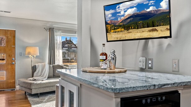 A modern kitchen with a marble countertop, a built-in dishwasher, and a mounted flat-screen TV displaying a scenic landscape. - Mountain Ave Retreat - Walk to Old Town Fun! (Fort Collins)