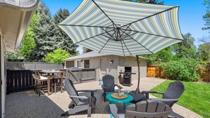 Une terrasse à l’arrière de la maison, avec des fauteuils Adirondack, un parasol à rayures, un barbecue et une table en bois.