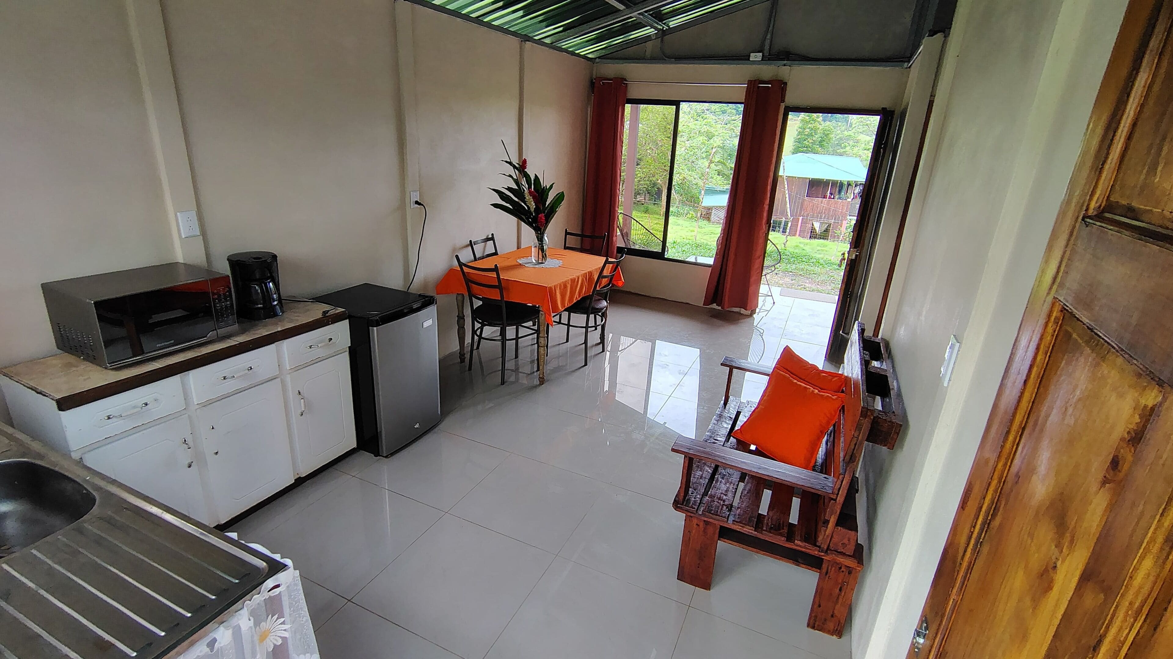 A compact kitchen with a microwave, coffee maker, and small refrigerator. There is a dining area with a table and chairs, and a view of the outdoors through a window.