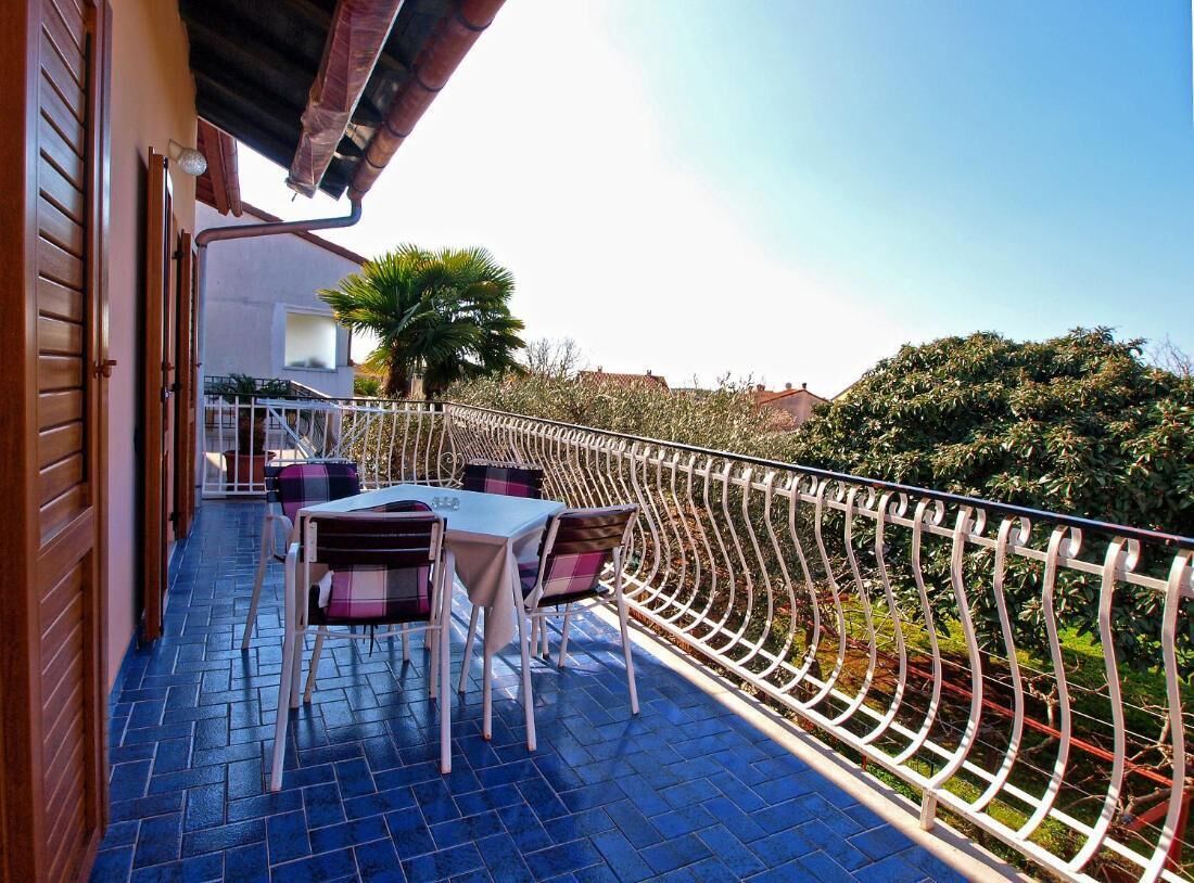 A balcony with a table and chairs, a white railing, and a view of greenery and buildings.
