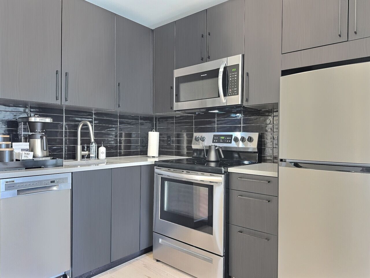 A modern kitchen with stainless steel appliances, grey cabinetry, and black tiled backsplash.