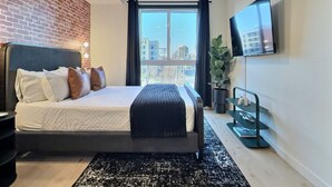 Ein modernes Schlafzimmer mit einem großen Bett, einem Deckenventilator, einem an der Wand montierten Fernseher und Blick auf die Stadt durch ein Fenster.