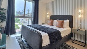 A modern bedroom with a large bed, a nightstand, a desk, and a view of the city through a large window.