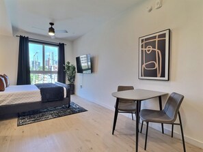 Une chambre d’hôtel moderne équipée d’un lit, d’une table à manger avec des chaises, d’un téléviseur fixé au mur et d’une grande fenêtre avec des rideaux.
