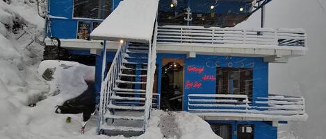 Un edificio de dos pisos con techo nevado y balcón, rodeado de nieve.