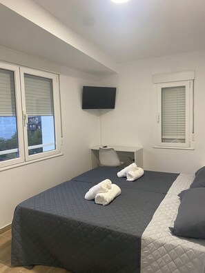 A neatly made bed with a dark quilt, two rolled towels, a flat-screen TV mounted on the wall, and a window with blinds. - Albergue O Bailarín (O Barco de Valdeorras)