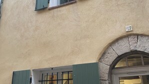 Un bâtiment à la façade beige, doté d’une petite fenêtre avec des volets verts et d’une entrée en arc de pierre.