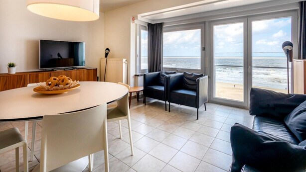 Ein modernes Wohnzimmer mit großem Fenster und Meerblick, einem Essbereich mit Tisch und Stühlen sowie einem Flachbildfernseher auf einer Holzkommode.