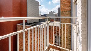 Ein Balkon mit weißer Brüstung, der auf ein Backsteingebäude mit Flachdach und Schornstein blickt.