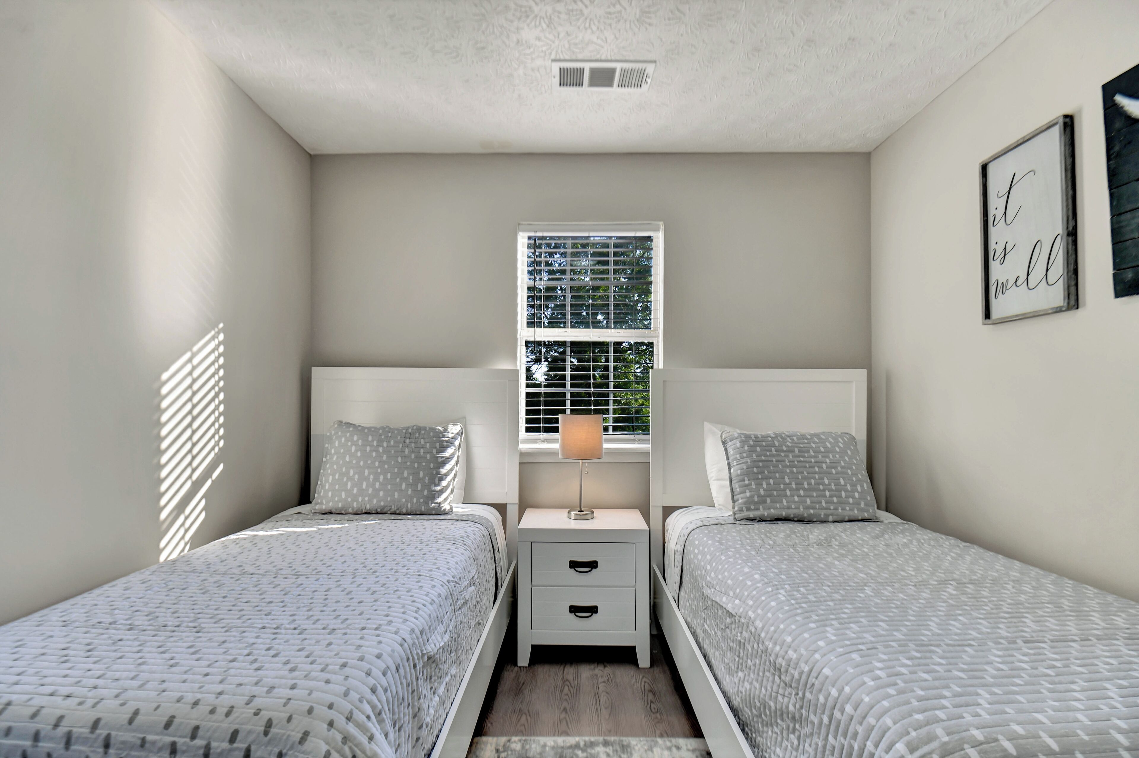 A bedroom with two beds, a nightstand with drawers, a window with blinds, and a wall-mounted sign.