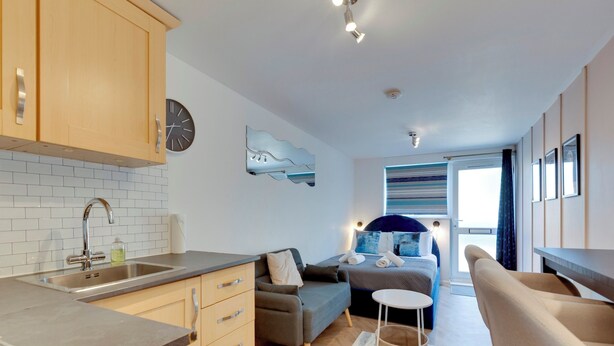 Una cocina moderna con un lavamanos, armarios y un reloj en la pared. Hay una zona de comedor con una mesa y sillas, y una sala de estar con un sofá y una pequeña mesa redonda.