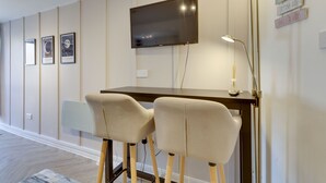 Una habitación moderna con una televisión de pantalla plana en la pared, dos taburetes tipo bar, un escritorio y una lámpara de pie.