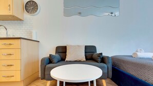 Una habitación compacta con kitchenette, un sofá con cojines, una mesa redonda blanca y una cama con manta.