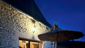 An outdoor patio area with a wooden table and benches, a thatched umbrella, and a stone building with a glass door leading to an interior space. - Charming gîte with swimming pool (Rouffignac-Saint-Cernin-de-Reilhac)