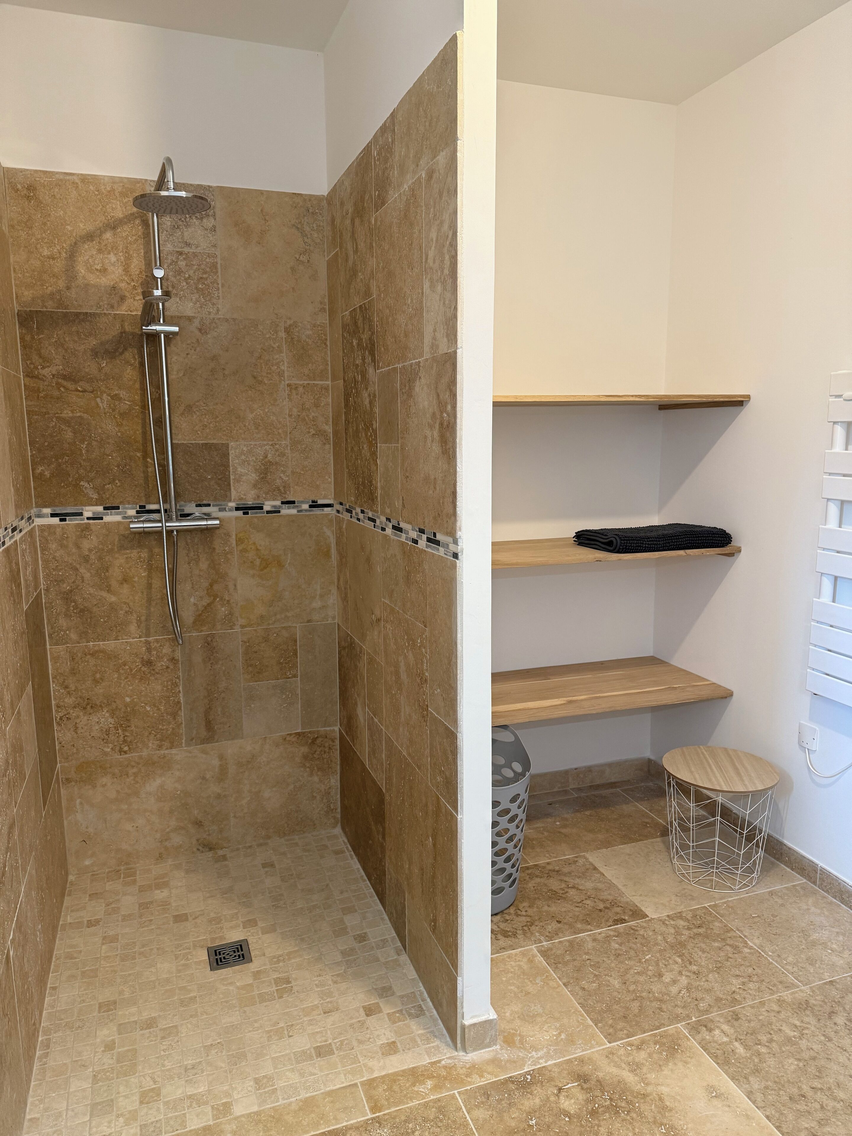 A bathroom with a shower area featuring beige tiles, a wooden shelf, and a small round table.