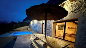 A pool area with a wooden deck and an umbrella, a stone wall, and a glass door leading to a room with a sofa and a dining table. - Charming gîte with swimming pool (Rouffignac-Saint-Cernin-de-Reilhac)