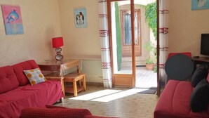 Een woonkamer met een rode bank, een glazen salontafel en een televisie. Er staat een klein houten tafeltje met een rode lamp en een stoel. De kamer heeft een schuifdeur van glas die uitkomt op een balkon met een bloempot.