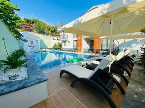 A pool area with lounge chairs and umbrellas, surrounded by greenery and a building with a balcony. - Nurhal (Ortaca)