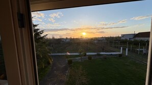 Vista a través de una ventana que muestra una zona residencial con cielo despejado, un atardecer y un camino pavimentado que conduce a un área verde con una valla y algunos árboles.