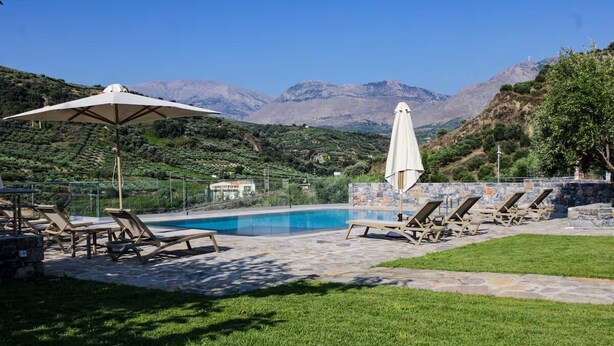 Area piscina con lettini e ombrelloni, circondata da un muro in pietra e rigogliosa vegetazione, con montagne sullo sfondo.