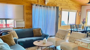 Een gezellig chaletinterieur met houten plafond, een opgehangen hertenkop, een blauwe bank, een ronde salontafel en een haard van baksteen.