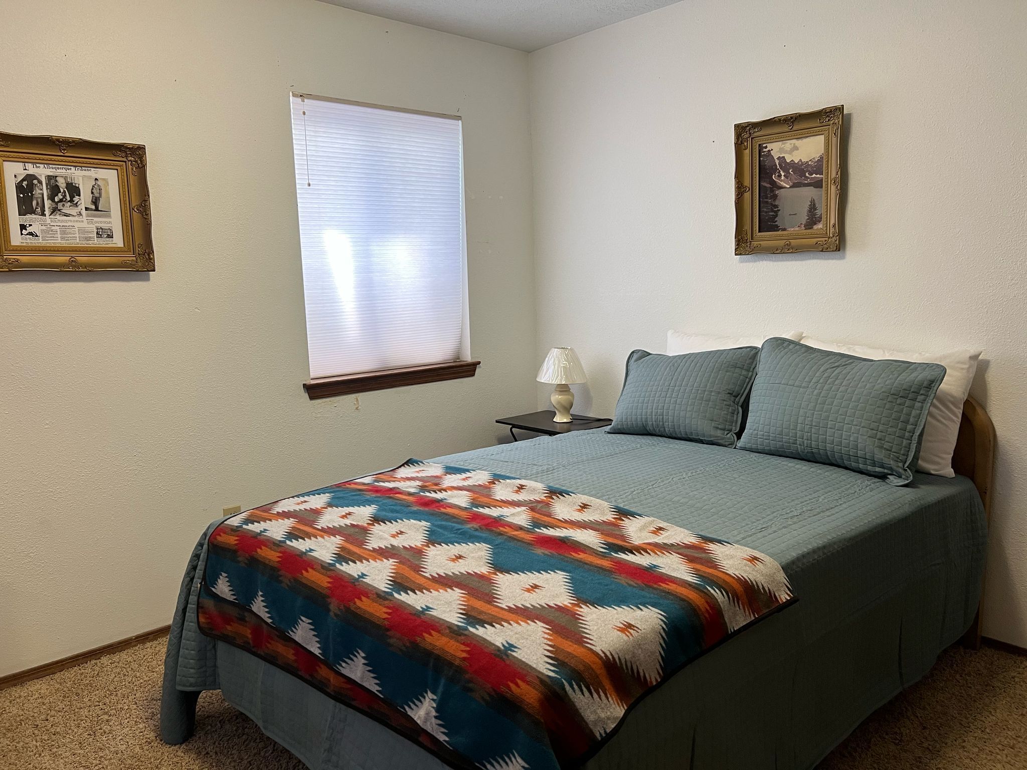 Une chambre à coucher avec un lit, une fenêtre avec des stores, une table de chevet avec une lampe et deux tableaux encadrés accrochés au mur.