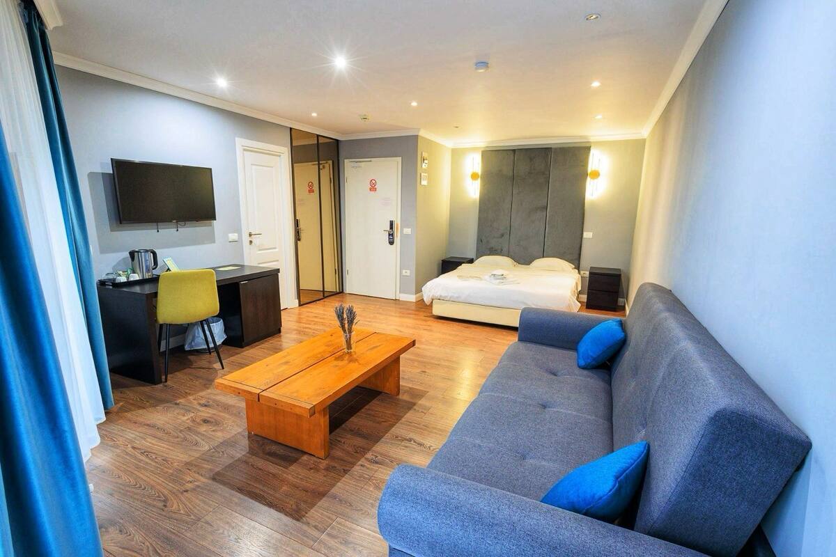 Una habitación de hotel moderna con suelo de madera, un sofá con cojines azules, una cama, un escritorio con una silla, un televisor montado en la pared y una puerta con un cartel.