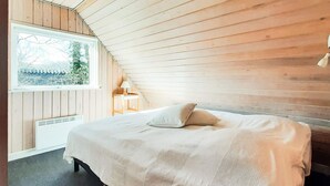 Ein Schlafzimmer mit schrägem Dach, einem Bett mit weißer Bettdecke, einem Nachttisch mit Lampe und einem Fenster mit Blick auf Bäume.