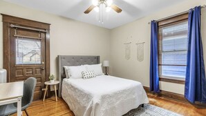 Une chambre à coucher comprenant un lit, un bureau, une chaise, une petite table avec une plante, un ventilateur de plafond et une fenêtre avec des rideaux.