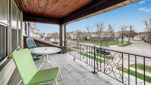 Un balcon doté d’un plafond en bois, de chaises vertes, d’une petite table et offrant une vue sur une rue de banlieue.