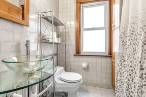 Een badkamer met een wasbak van glas, een toilet, een handdoekhouder en een raam.