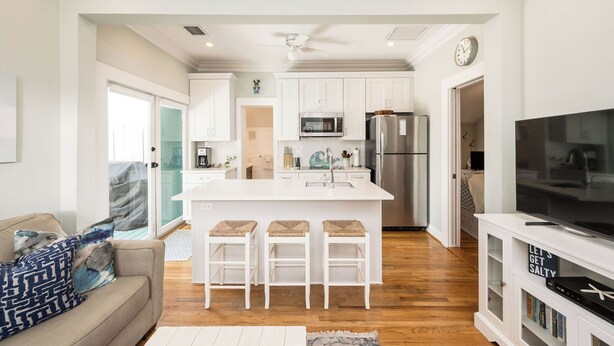 Una cocina moderna con armarios blancos, una isla central con taburetes, un microondas, un refrigerador y un televisor sobre un mueble blanco.