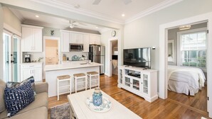 Una cocina moderna con armarios blancos, un microondas y un refrigerador. Hay una zona de comedor con una mesa y sillas blancas. Un televisor está montado en la pared sobre la zona de estar.