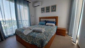 A bedroom with a bed, a wooden nightstand, a window with curtains, and a wall-mounted air conditioner.